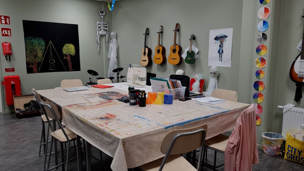 Ett rum med ett kvadratiskt bord med stolar. Bordet har fläckig duk. På bordet står bland annat en symaskin. På väggen hänger gitarrer och målningar.