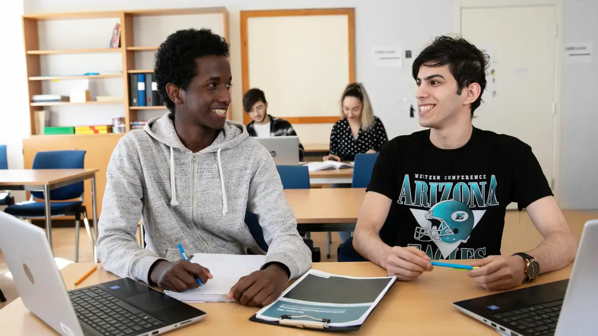 Två elever med uppslagna datorer diskuterar i klassrummet. I bakgrunden två andra elever.
