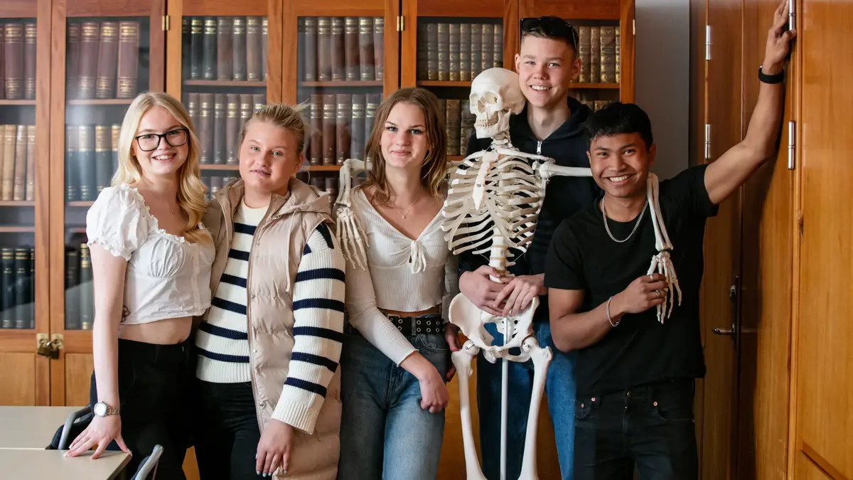 Fem elever och en skelettmodell poserar framför ett skåp fullt av böcker.
