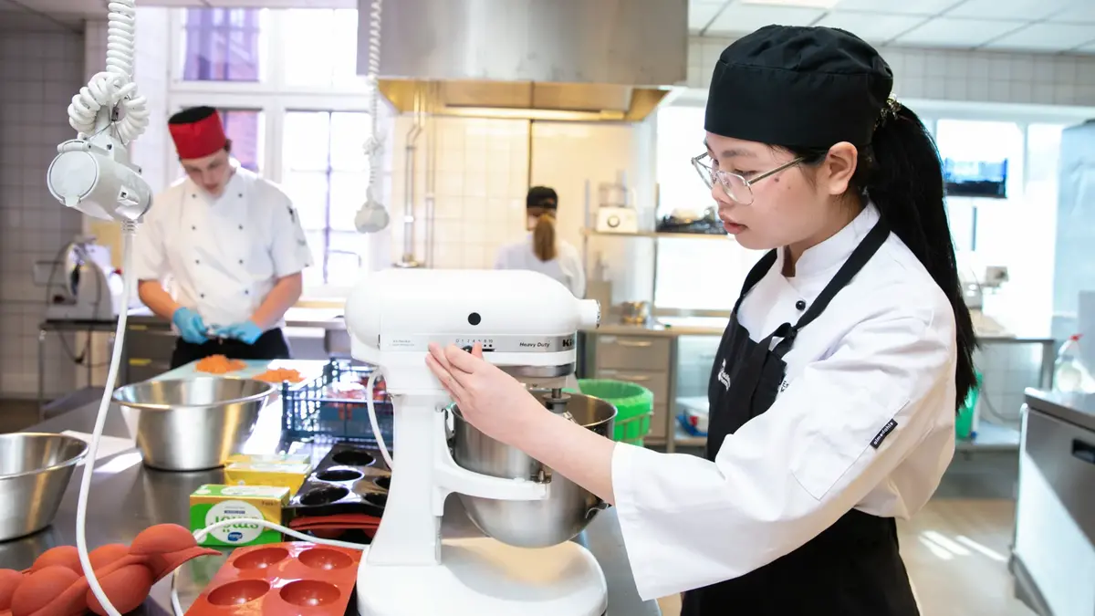 Elev iklädd förkläde står i köket med en köksmaskin. I bakgrunden en annan elev som står och lagar mat.