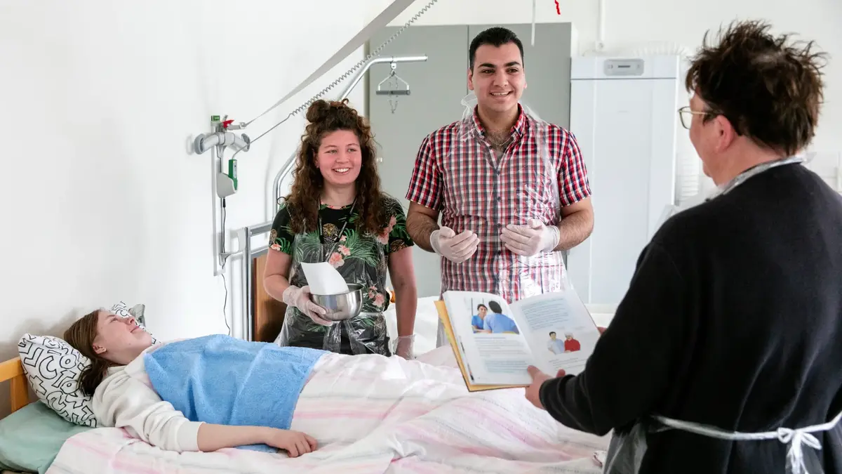 Lärare instruerar två tre elever i vårdkunskap. En elev ligger i en sjuksäng och två elever står bredvid.